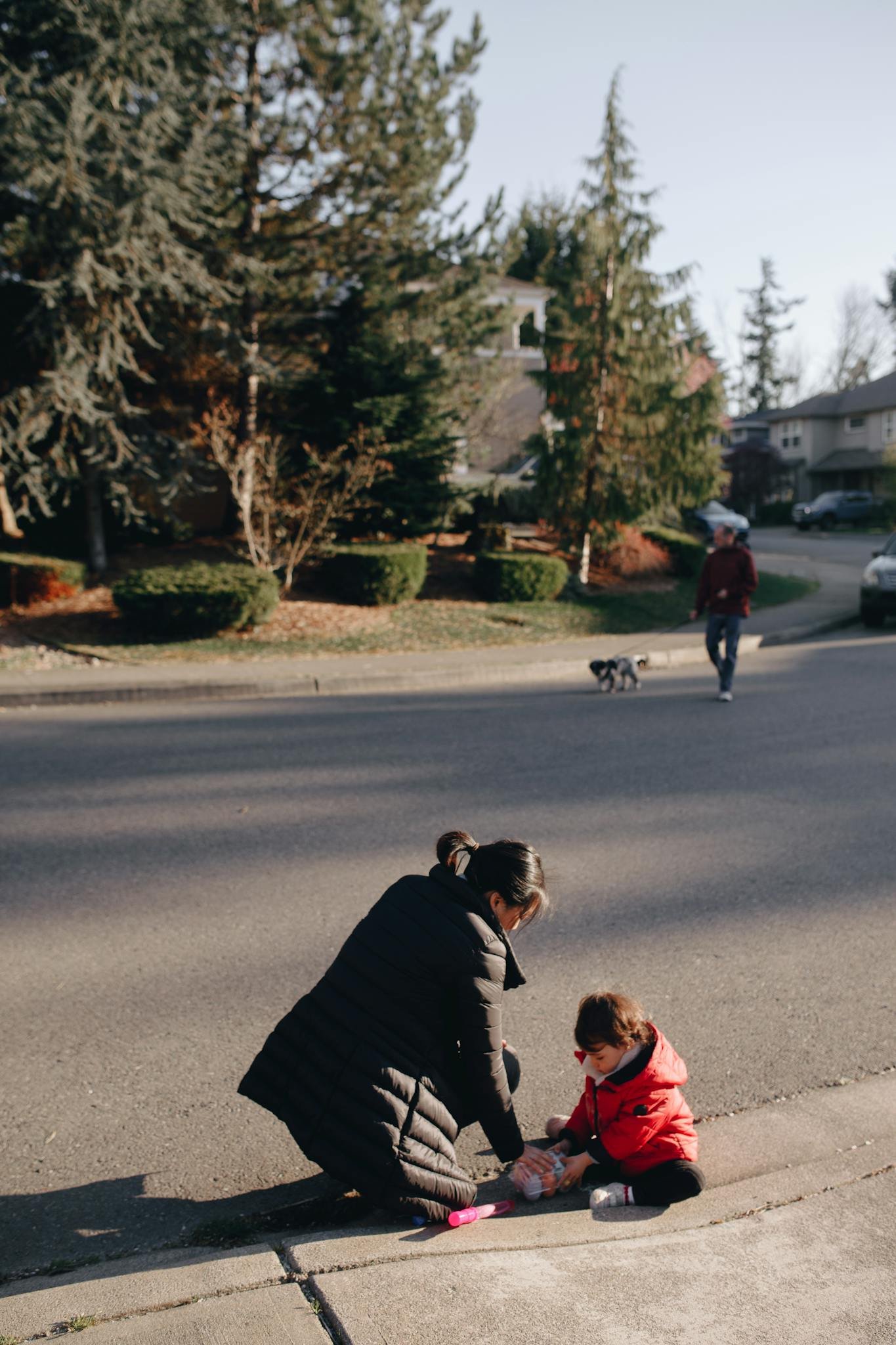 A mother helps her child on the sidewalk while a man walks a dog nearby.